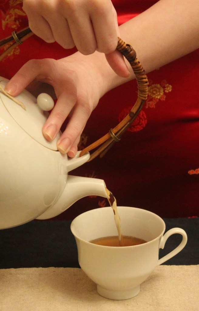 A photograph of a light skinned person pouring tea from a white teapot into a matching teacup. The background is red.
